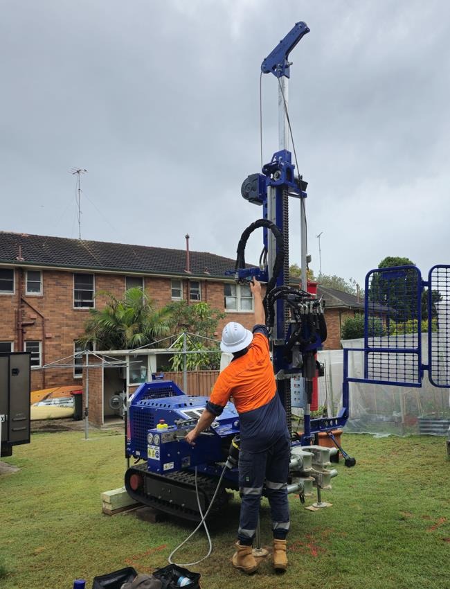 Blue tracked mini rig set up for drilling on a residential back lawn with an operator at the controls